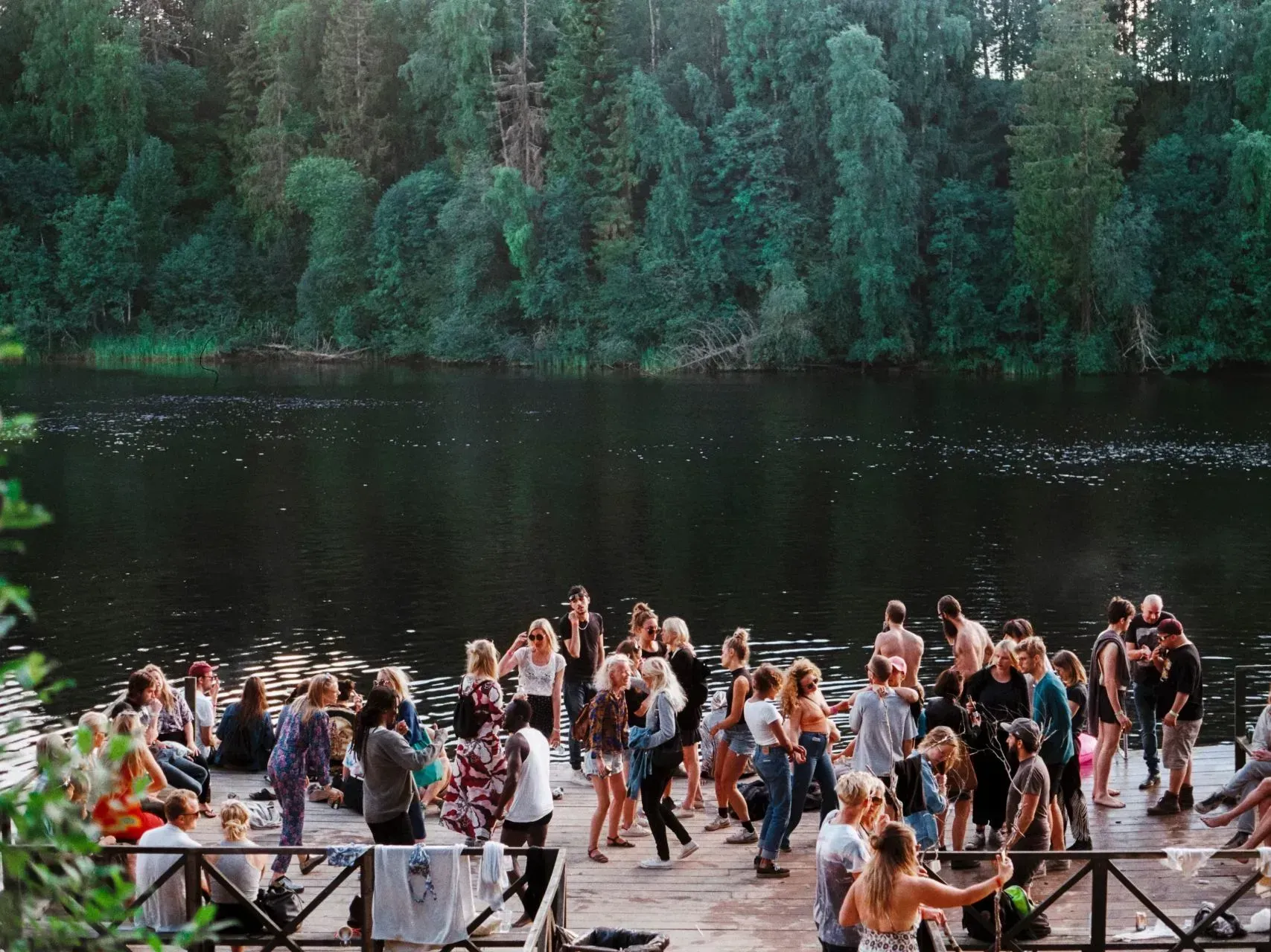 Folk på en trebrygge ved en innsjø, dansende. Skog i bakgrunnen. Kveldslys.
