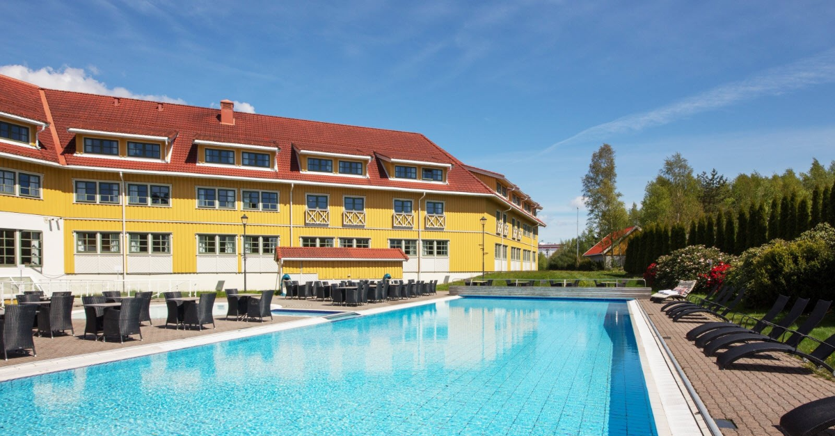 Hotell med gul fasade og rødt tak, utendørsbasseng, solrik dag.