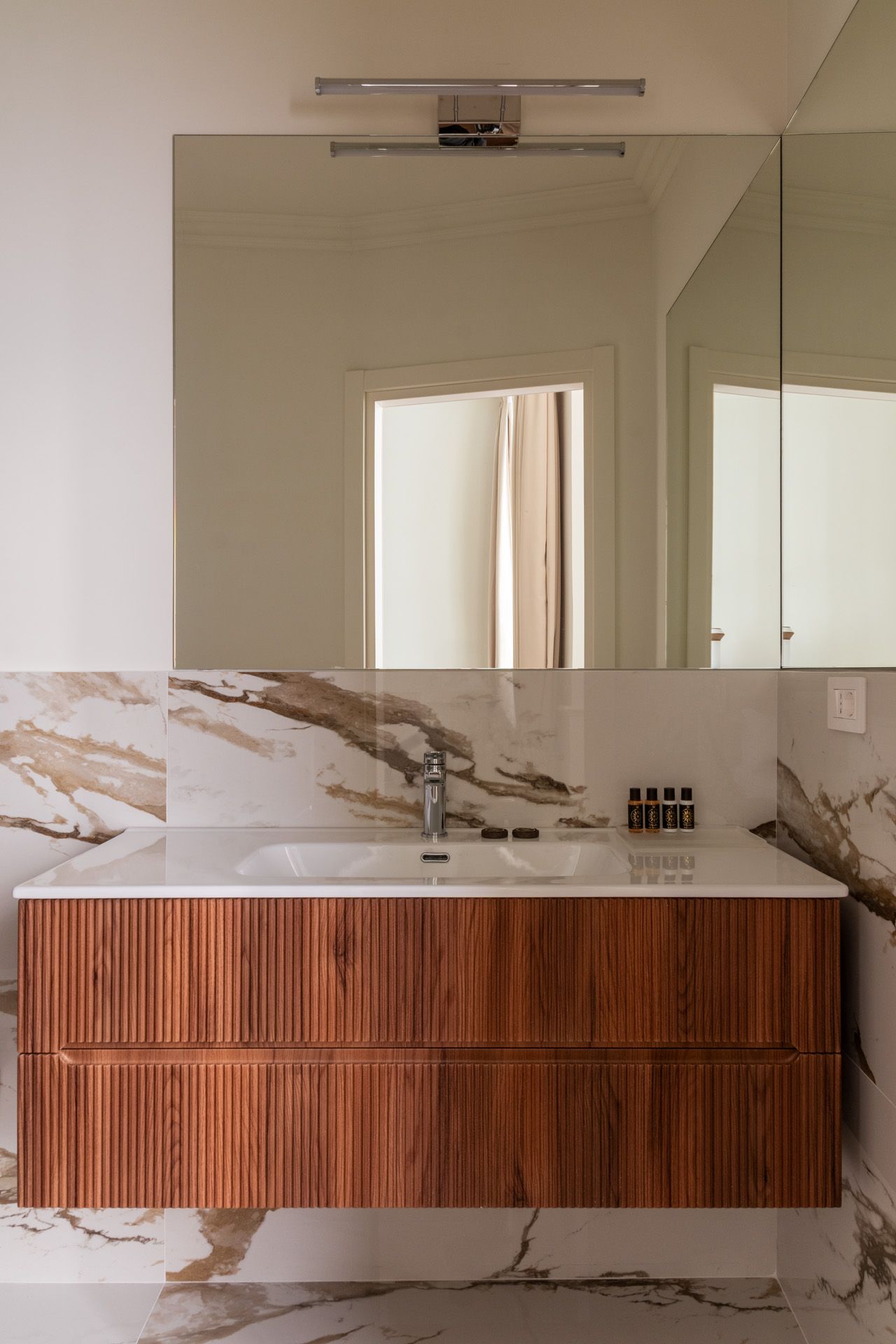 Wooden vanity with sink, beneath a large mirror and marble-patterned backsplash, with a chrome faucet.