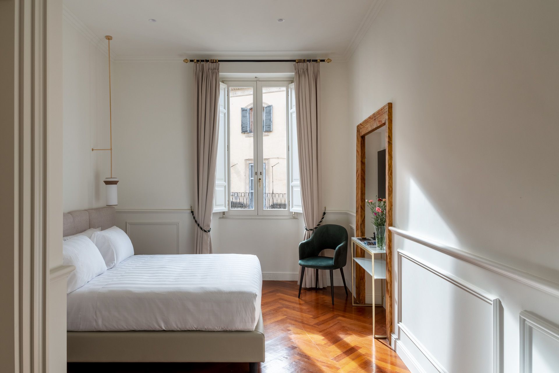 Bedroom with a bed, window, chair, and large mirror. Wooden floors and white walls.