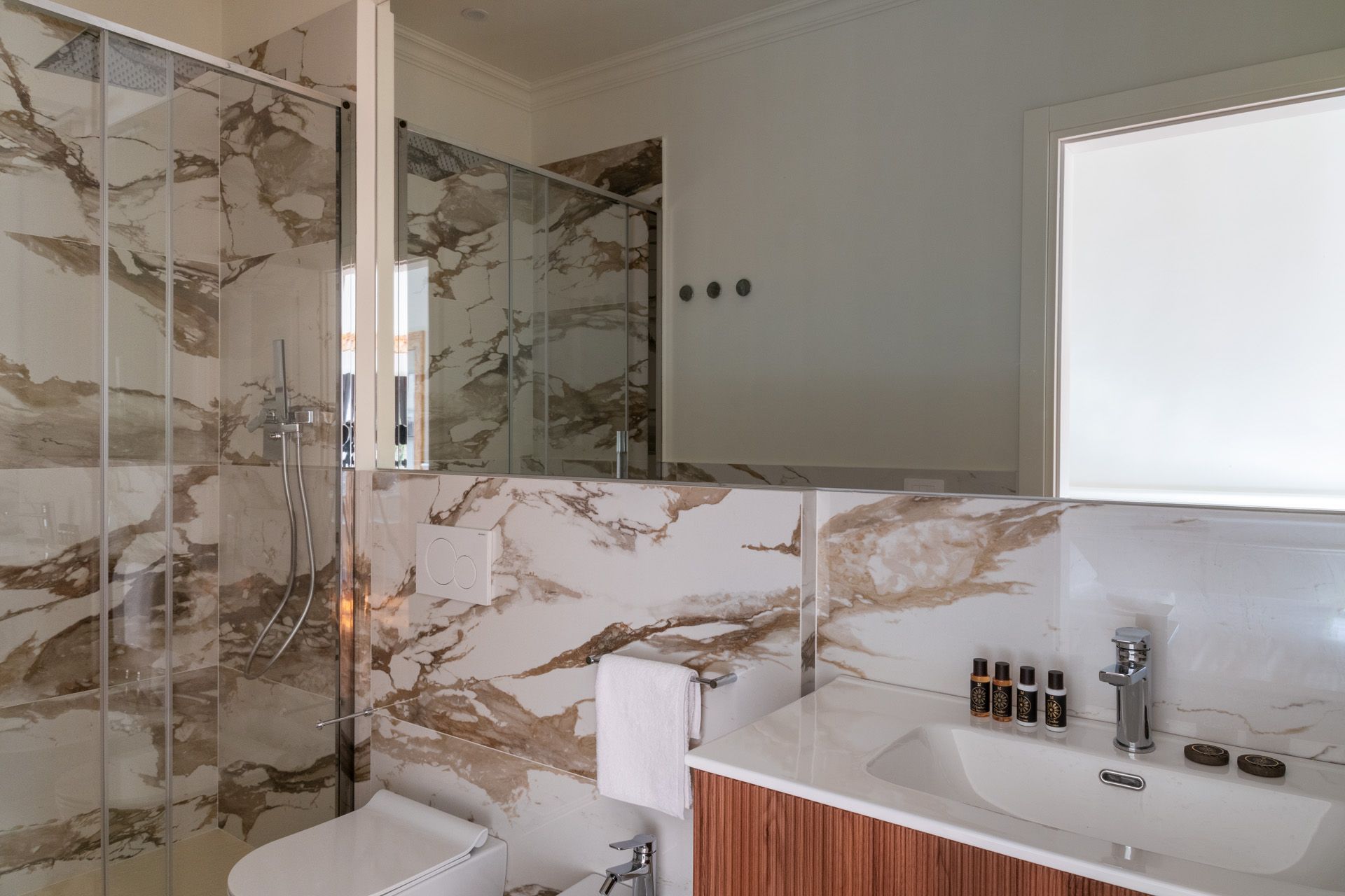 Bathroom with marble walls and glass shower. Vanity with sink, mirror, and toiletries.