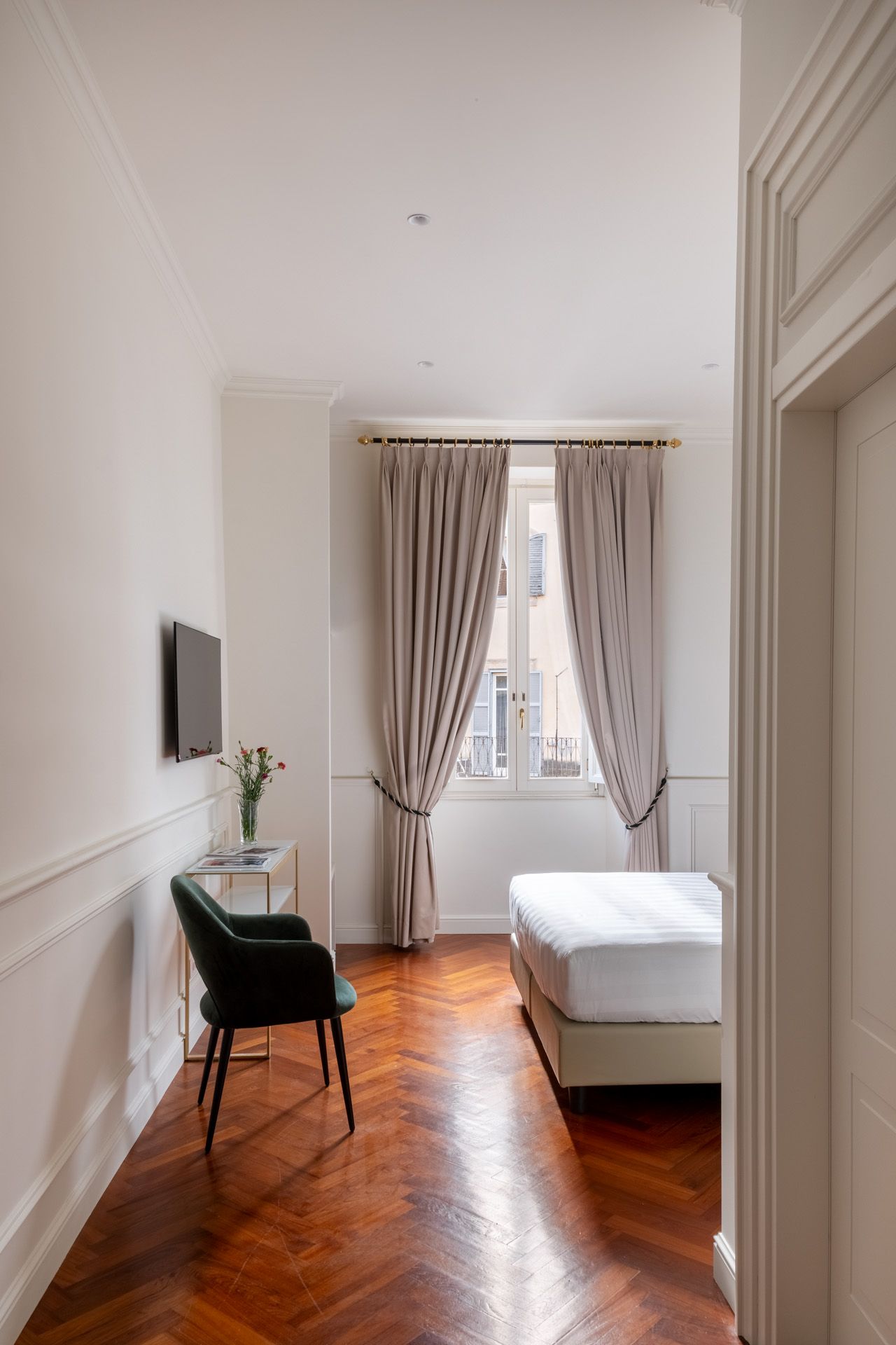 Bedroom with hardwood floor, bed by a window with curtains, chair, TV, and vase of flowers.