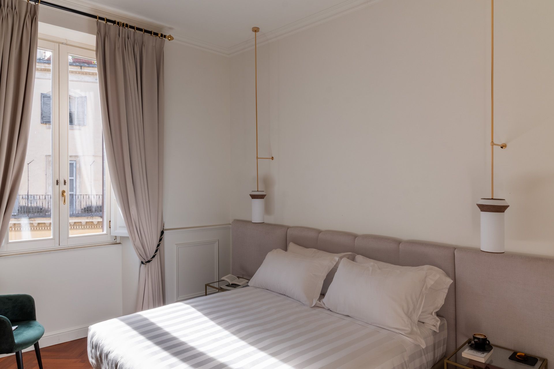 Bedroom with bed, pillows, window with drapes, and hanging lamps. Cream-colored walls, light wood trim, and a small chair.