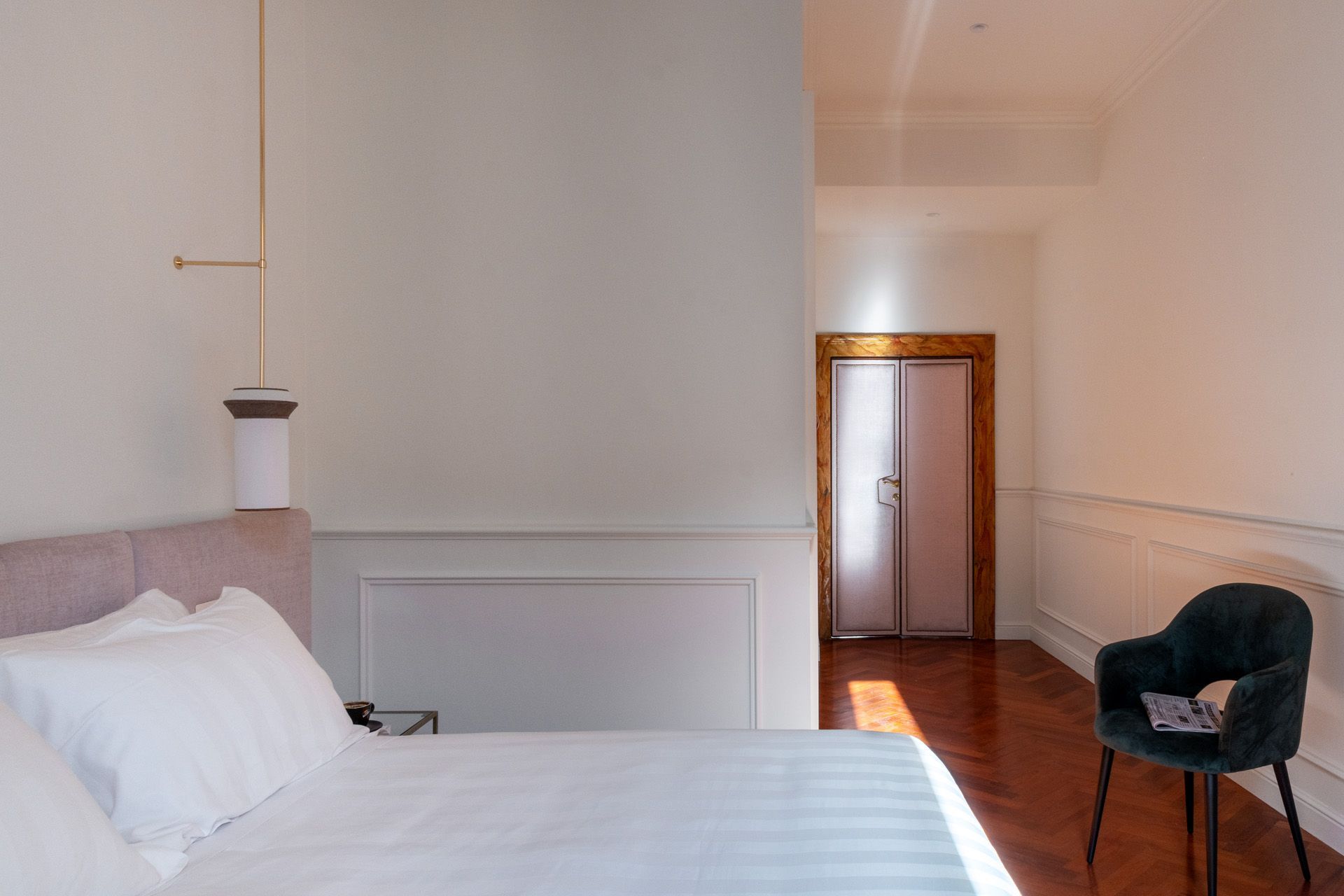 Bedroom with a bed, a chair, and a hallway leading to a closed door, bathed in sunlight.