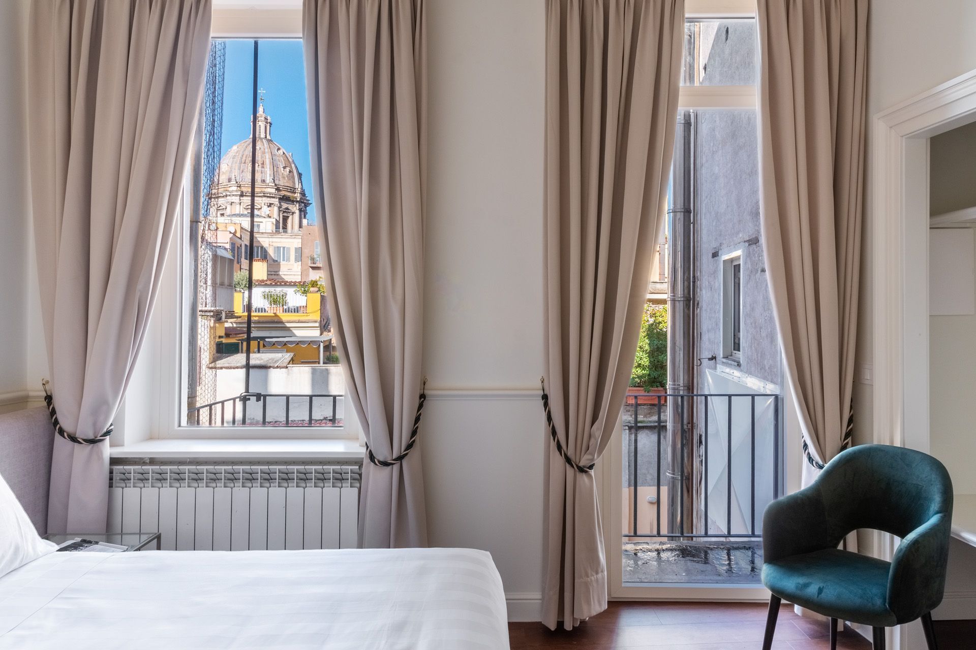 Bedroom with cityscape view; beige curtains frame windows, a blue chair sits near a door to a balcony.