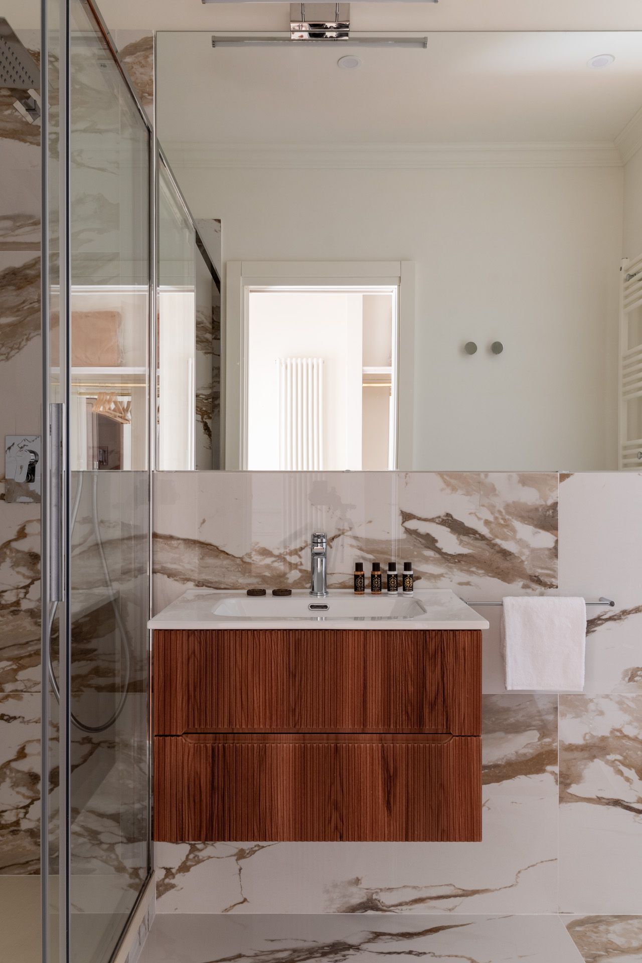 Bathroom with wood vanity, large mirror, and marble-patterned walls and floor. Glass shower.