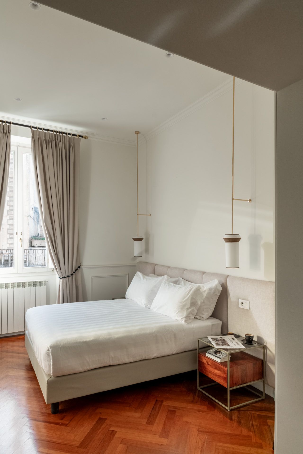 Bedroom with a bed, nightstand, window, and hanging lights. Features cream-colored walls, wood floors, and neutral curtains.