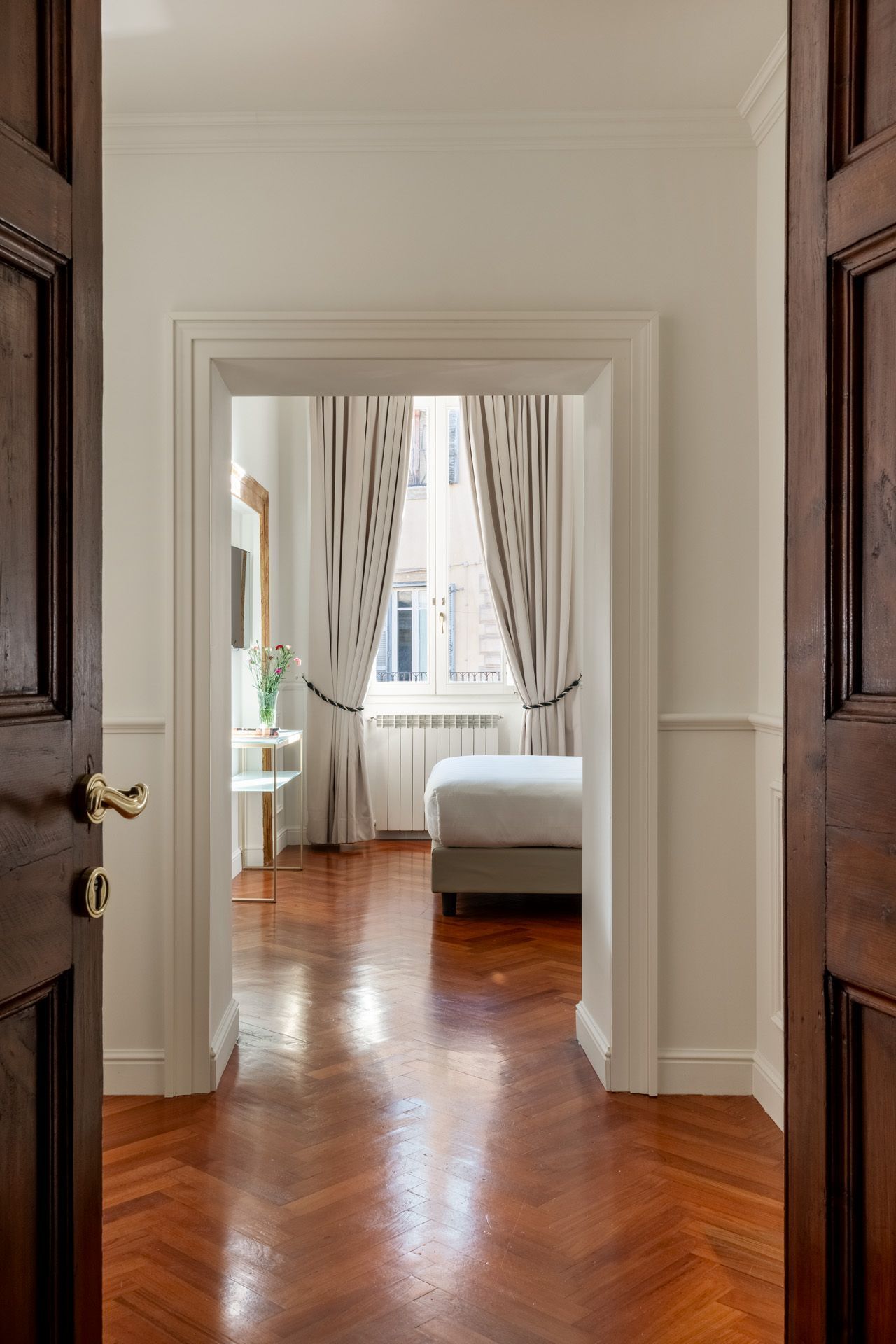 Open dark wooden doors reveal a hallway with a bedroom view. Hardwood floors, white walls, and a bed.