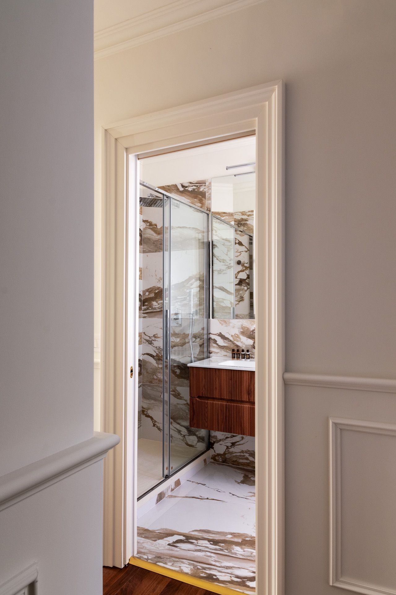 Hallway view into a bathroom with marble walls and a wooden vanity.