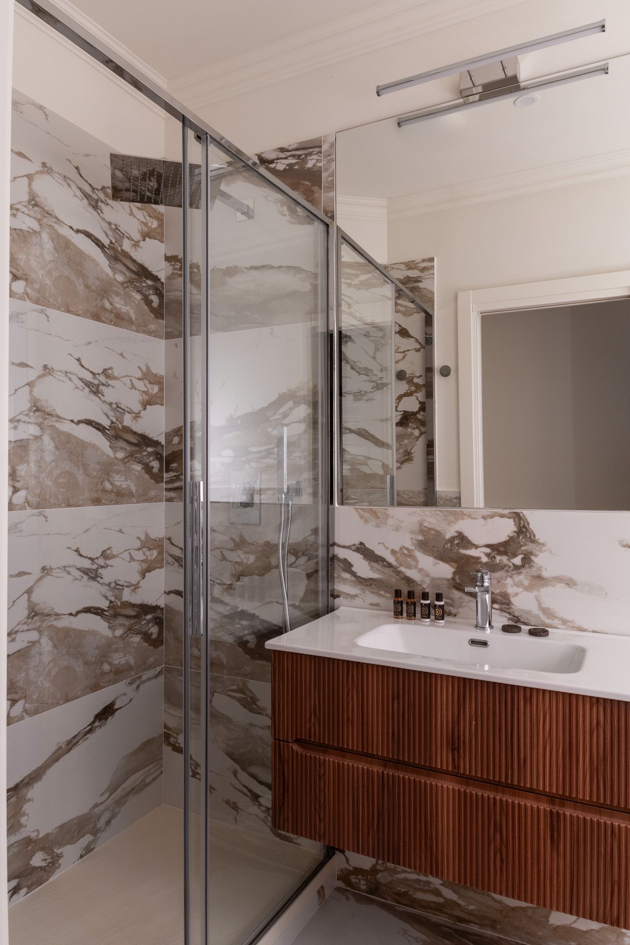 Bathroom with marble walls, a glass shower, a wood vanity, and a large mirror.