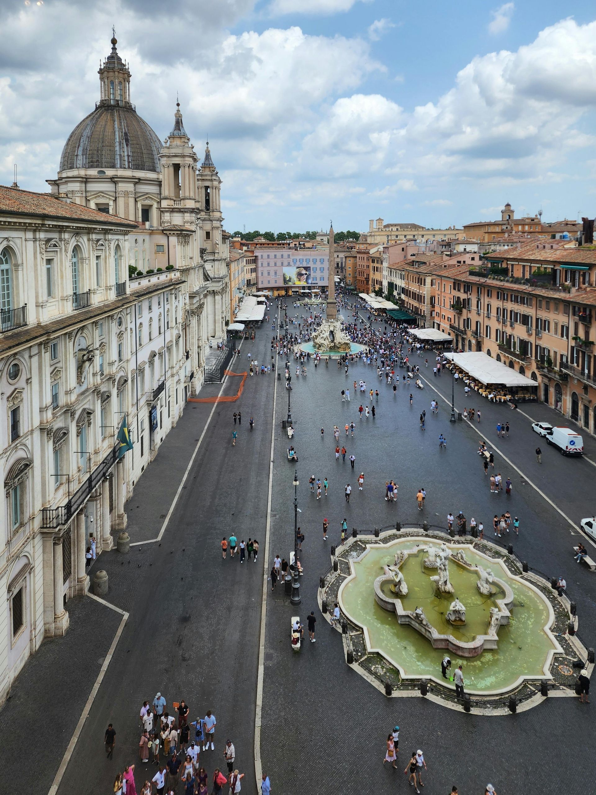 High-angle view of Piazza Navona, Rome, Italy, with people, fountains, and historic buildings under a cloudy sky.