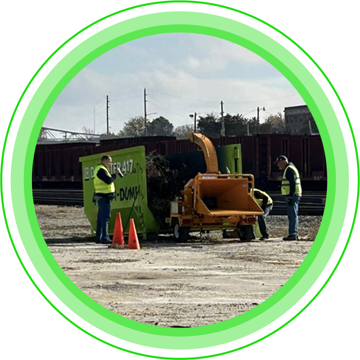 Men in safety vests operate a wood chipper near a green dumpster and train tracks.