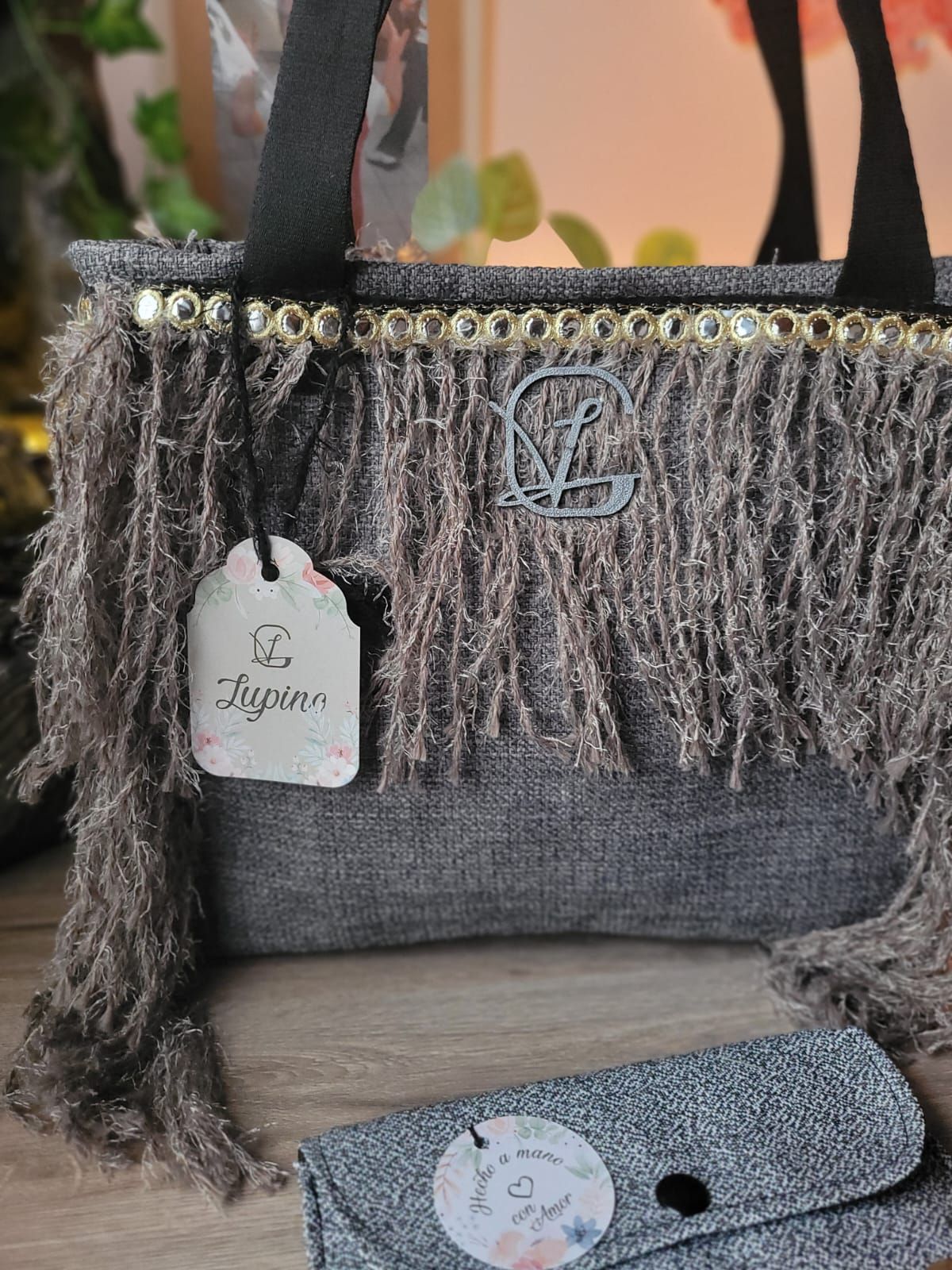 Un bolso gris con flecos y una etiqueta está sobre una mesa de madera.