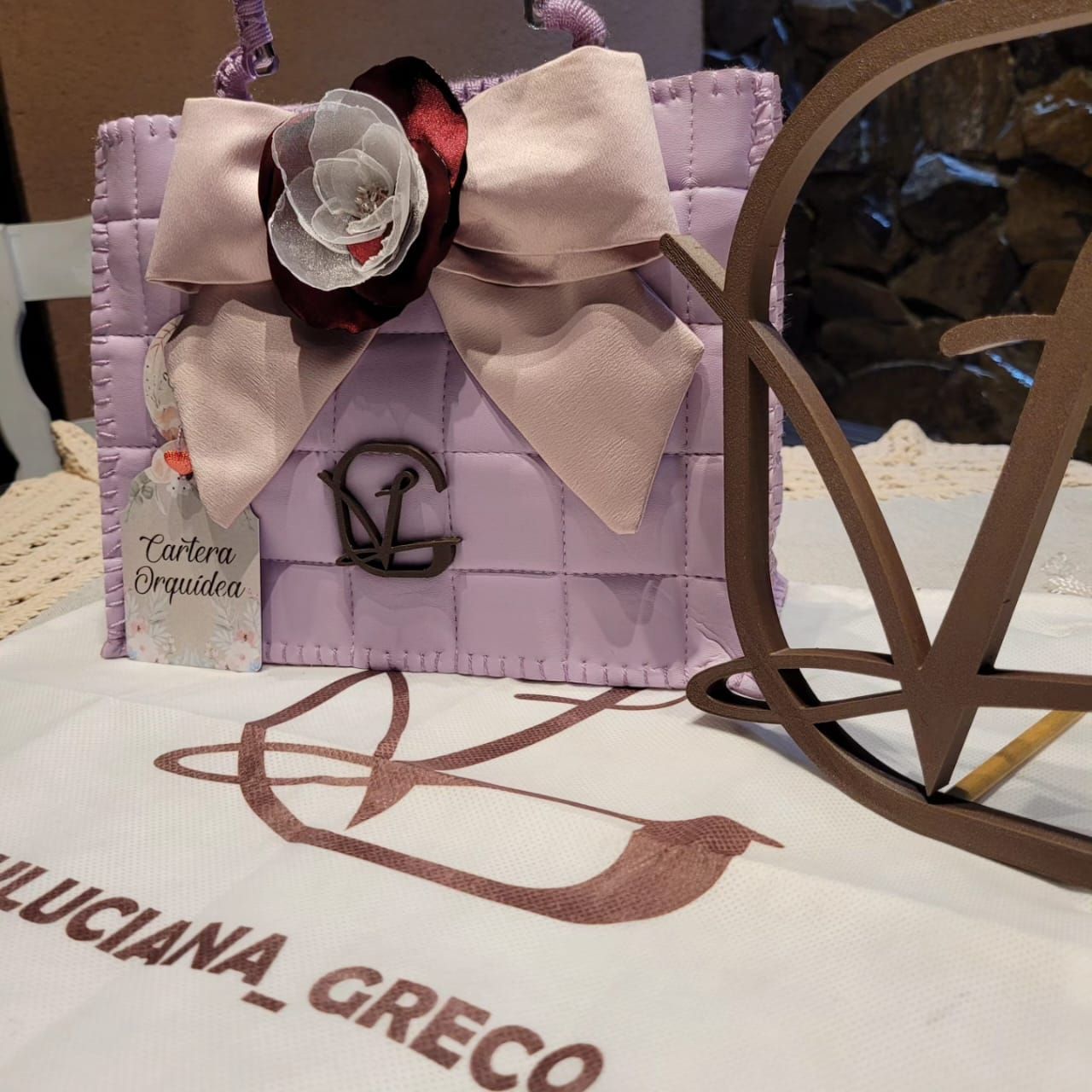 Un bolso morado reposa sobre una mesa junto a un cartel que dice Luciana Greco.