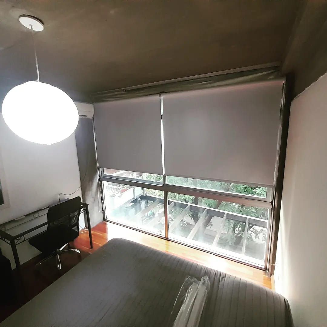 Un dormitorio con cama, escritorio y silla, y una gran ventana cubierta con estores enrollables de color claro.