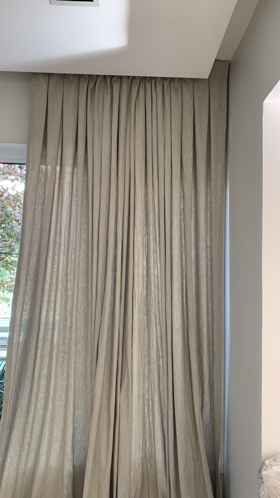 Cortinas beige que van del suelo al techo, colgadas en pliegues contra una pared y una ventana blancas.