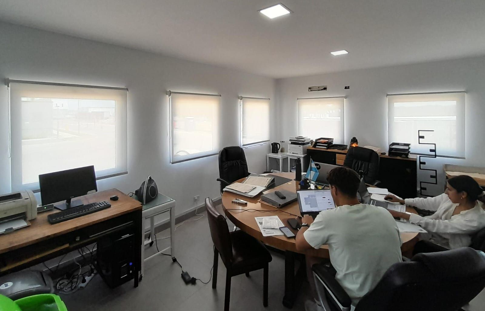 Ambiente de oficina con una mesa redonda de madera, dos personas trabajando y un escritorio aparte con equipo informático.