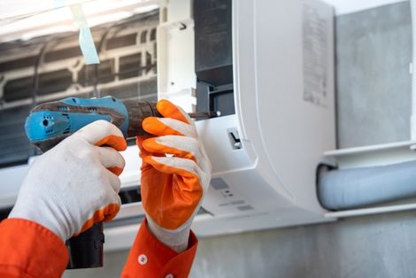 Technician drilling into a white wall-mounted air conditioner with orange and white gloves