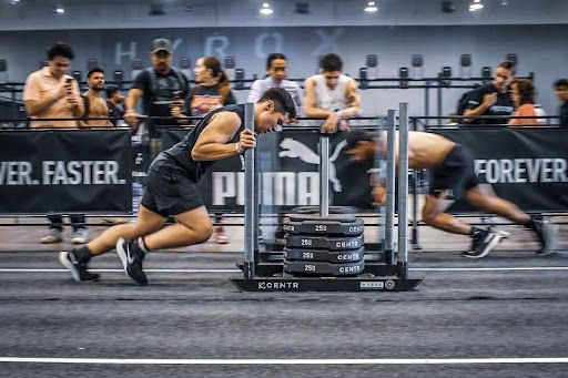 Two athletes push a weighted sled in a competition setting. They wear athletic gear, focused expressions, and move forward.