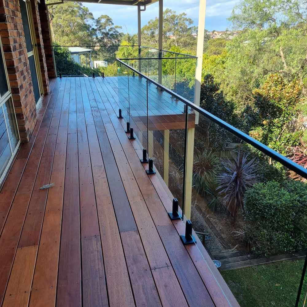 Wooden deck with a glass railing and trees in the background — Pool Fence in Unanderra NSW