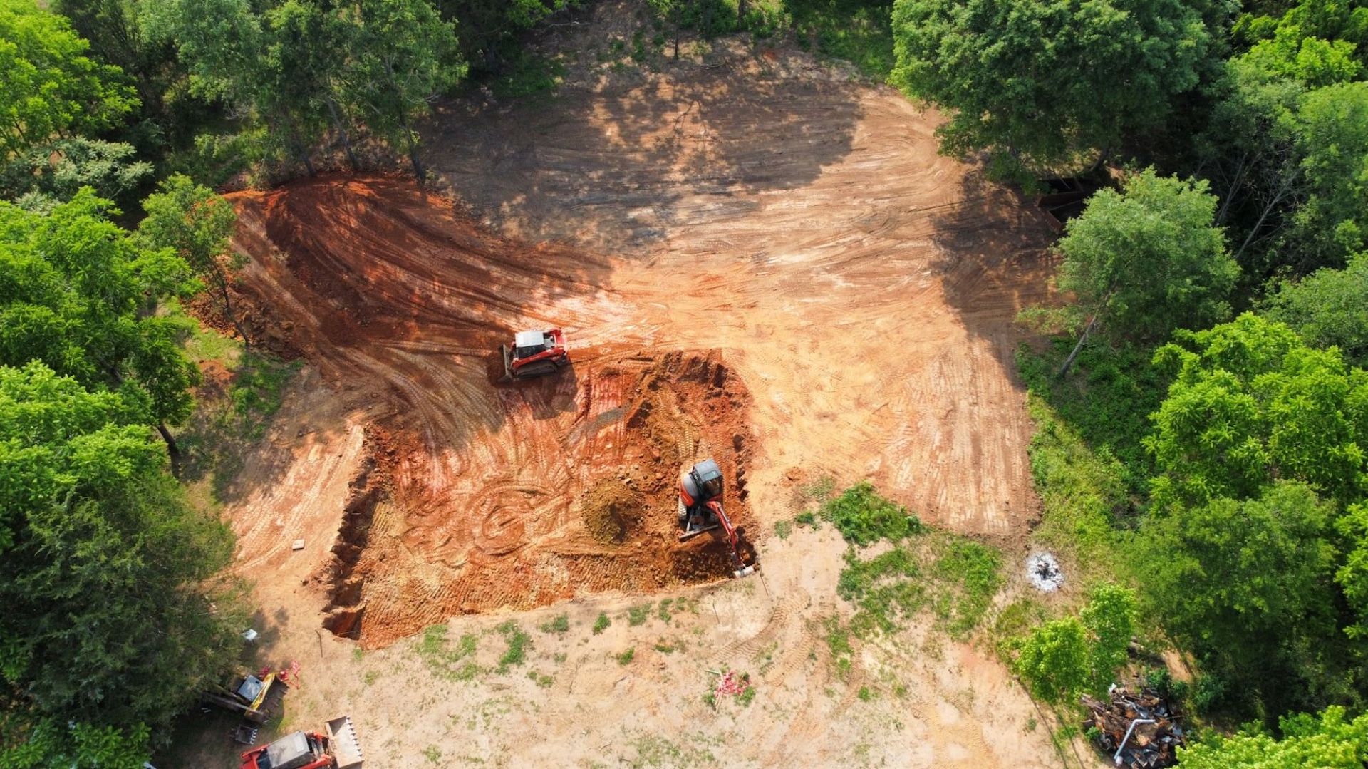 Aerial view of a cleared dirt plot in a wooded area with an excavator digging into the reddish-brown earth.