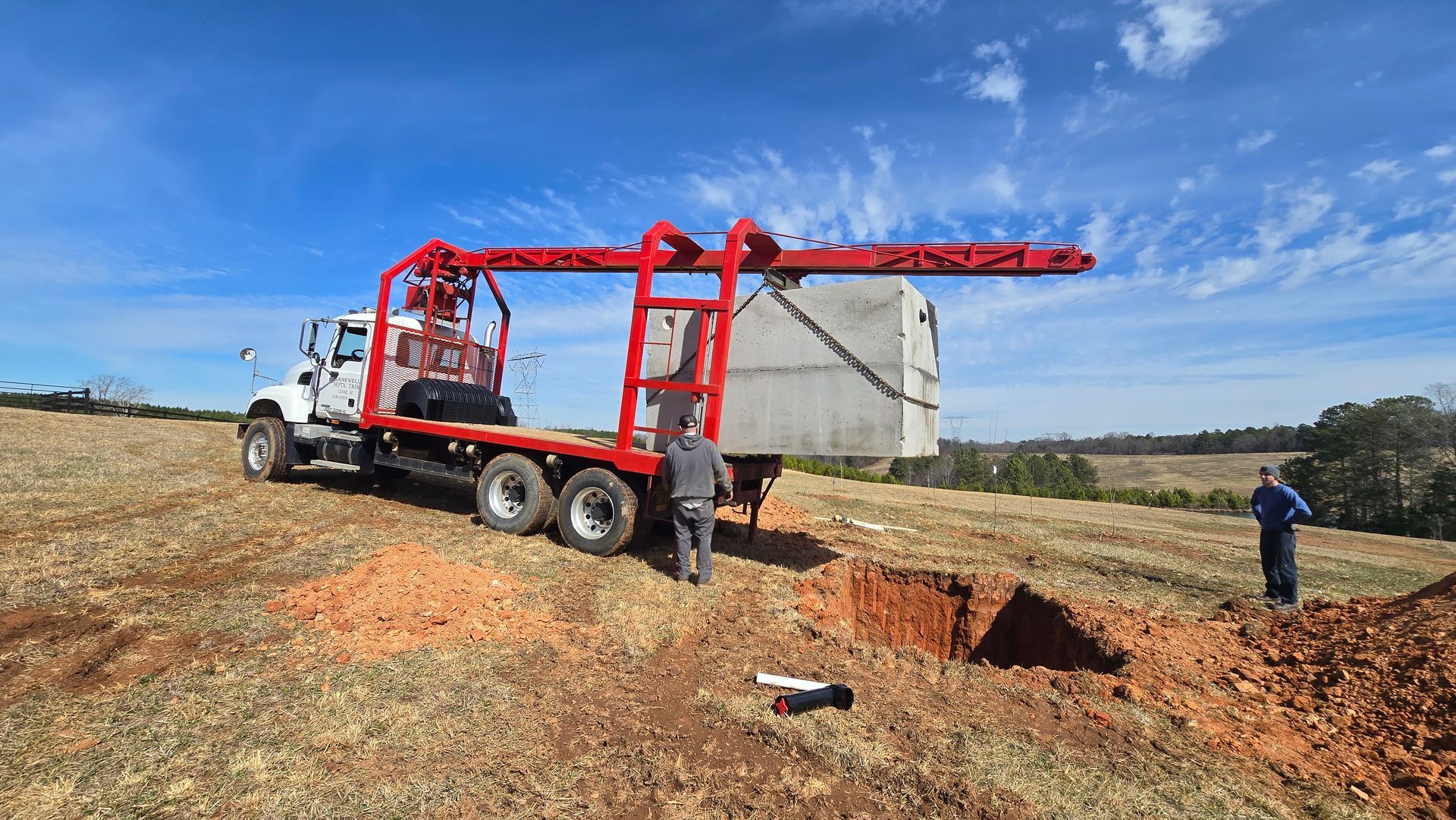 Truck-mounted crane lifting a large metal container in a rural field, with a worker nearby.