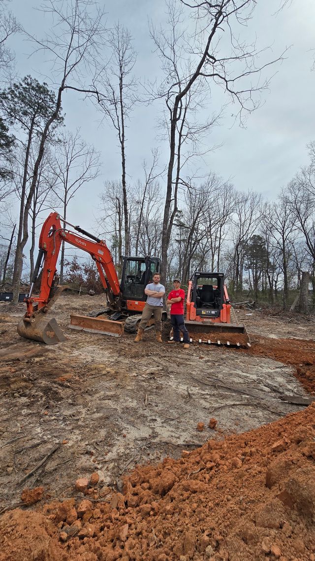 Two people stand between an orange excavator and a skid steer on a cleared dirt construction site surrounded by trees.