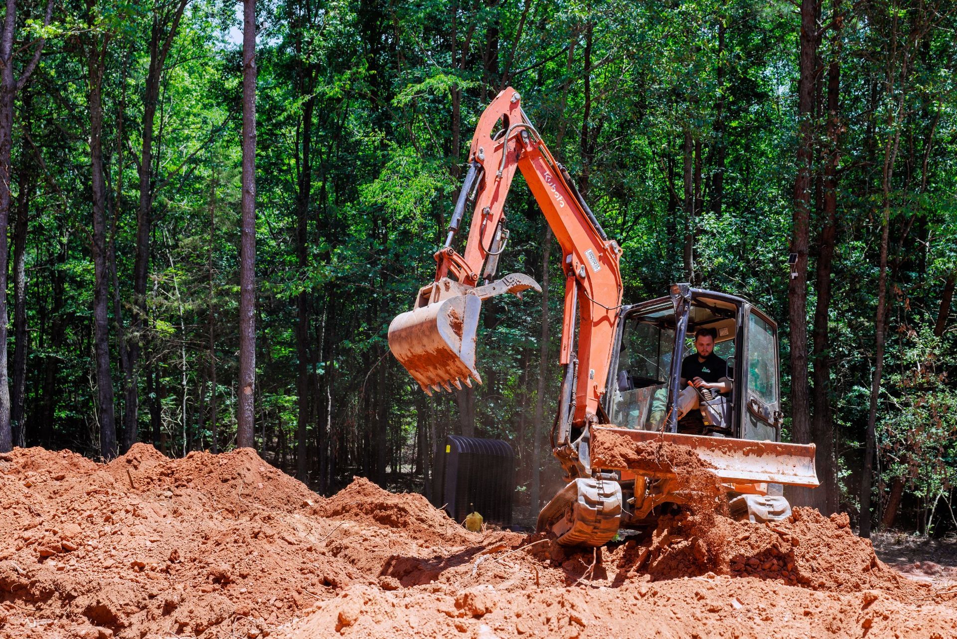 Orange excavator digging dirt in a forest clearing with tall trees
