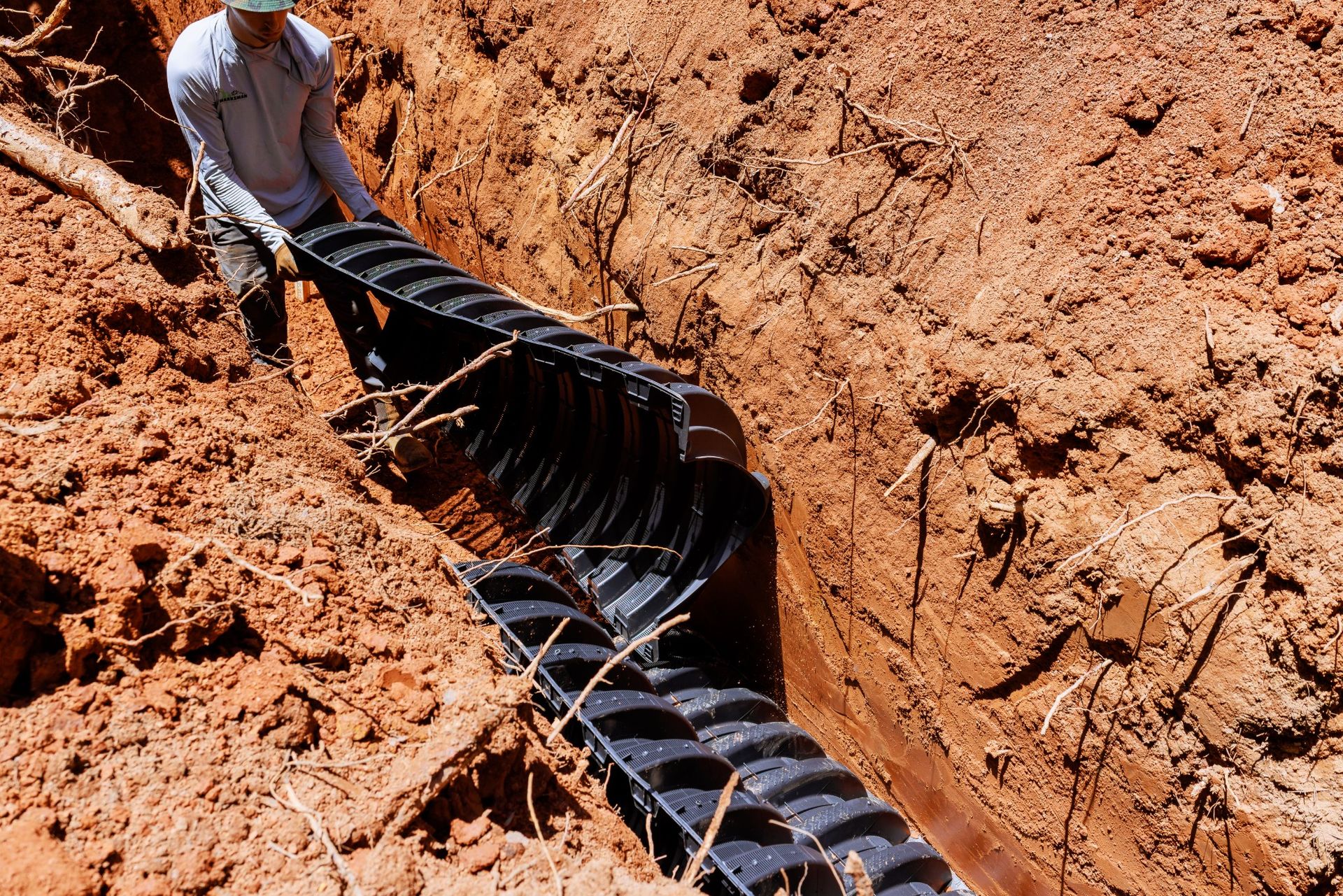 Worker installing a black corrugated drainage pipe in a red dirt trench
