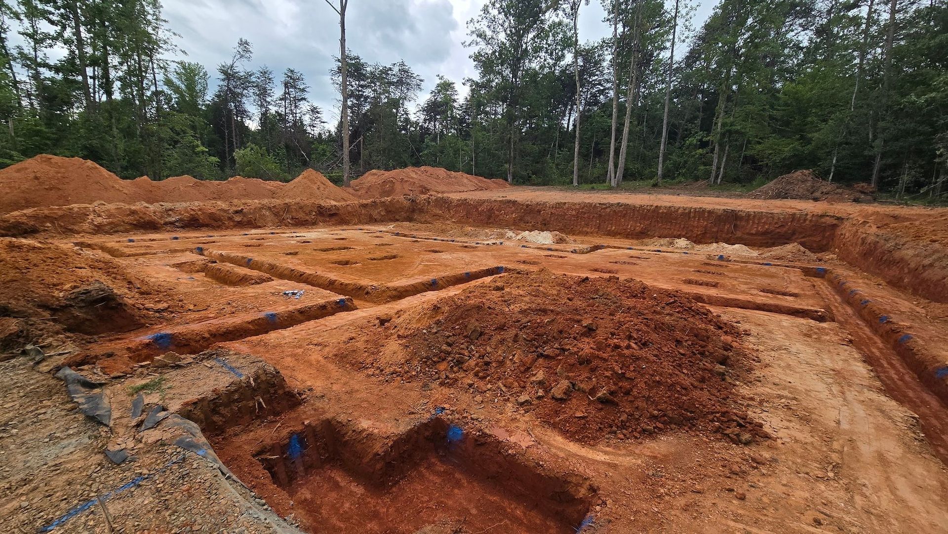 An excavated building site with trenches cut into red soil, surrounded by a forest under a cloudy sky.