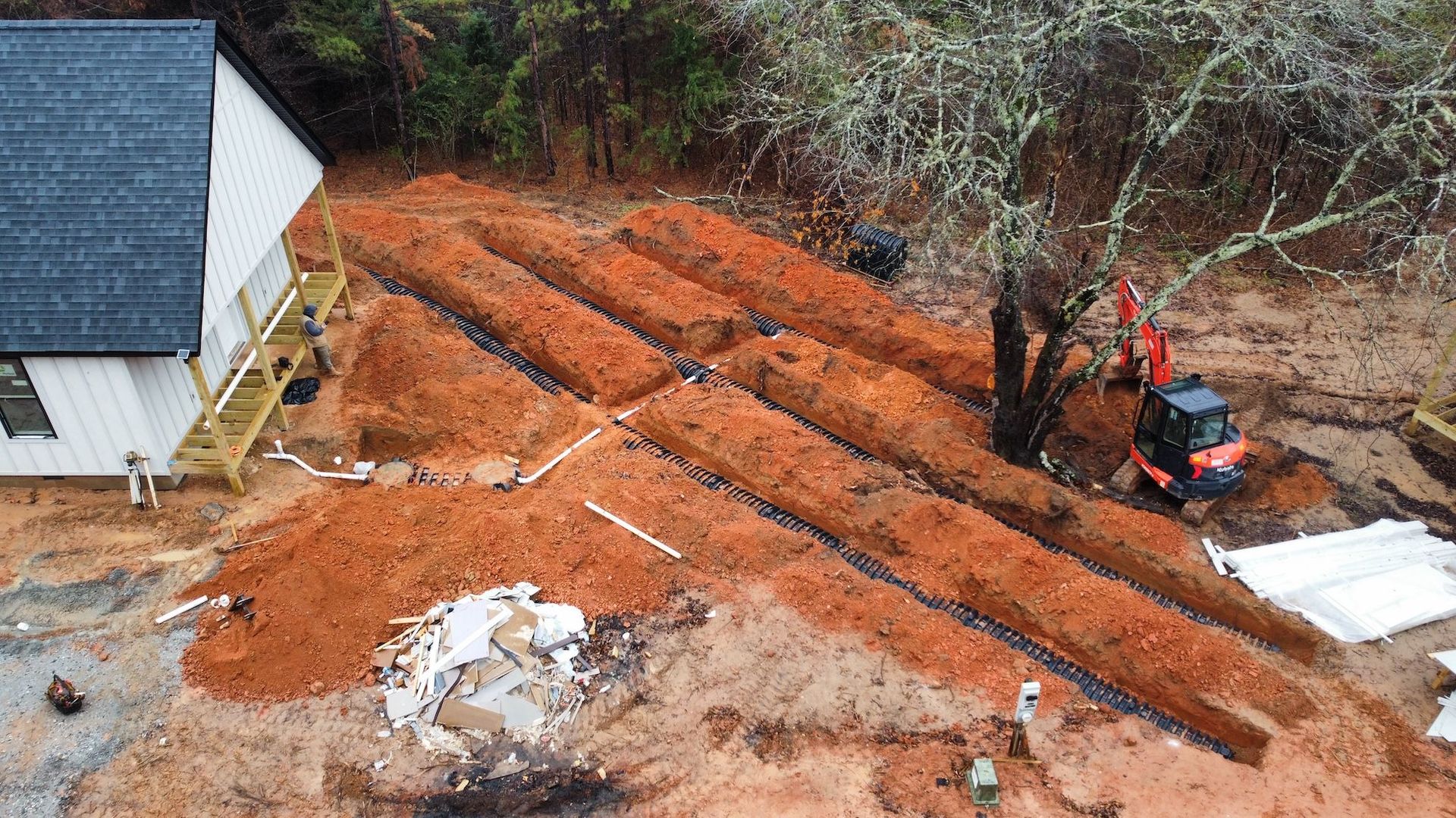 An aerial view shows an excavator digging long, parallel trenches in red dirt next to a house under construction.