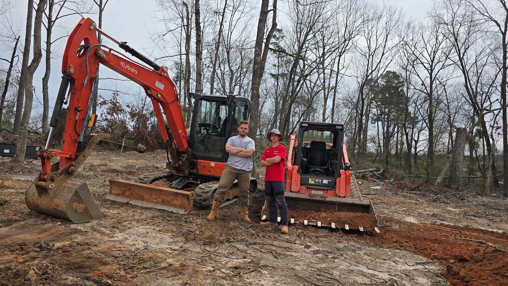 Two people stand between an orange excavator and a skid steer loader in a wooded, cleared dirt construction site.