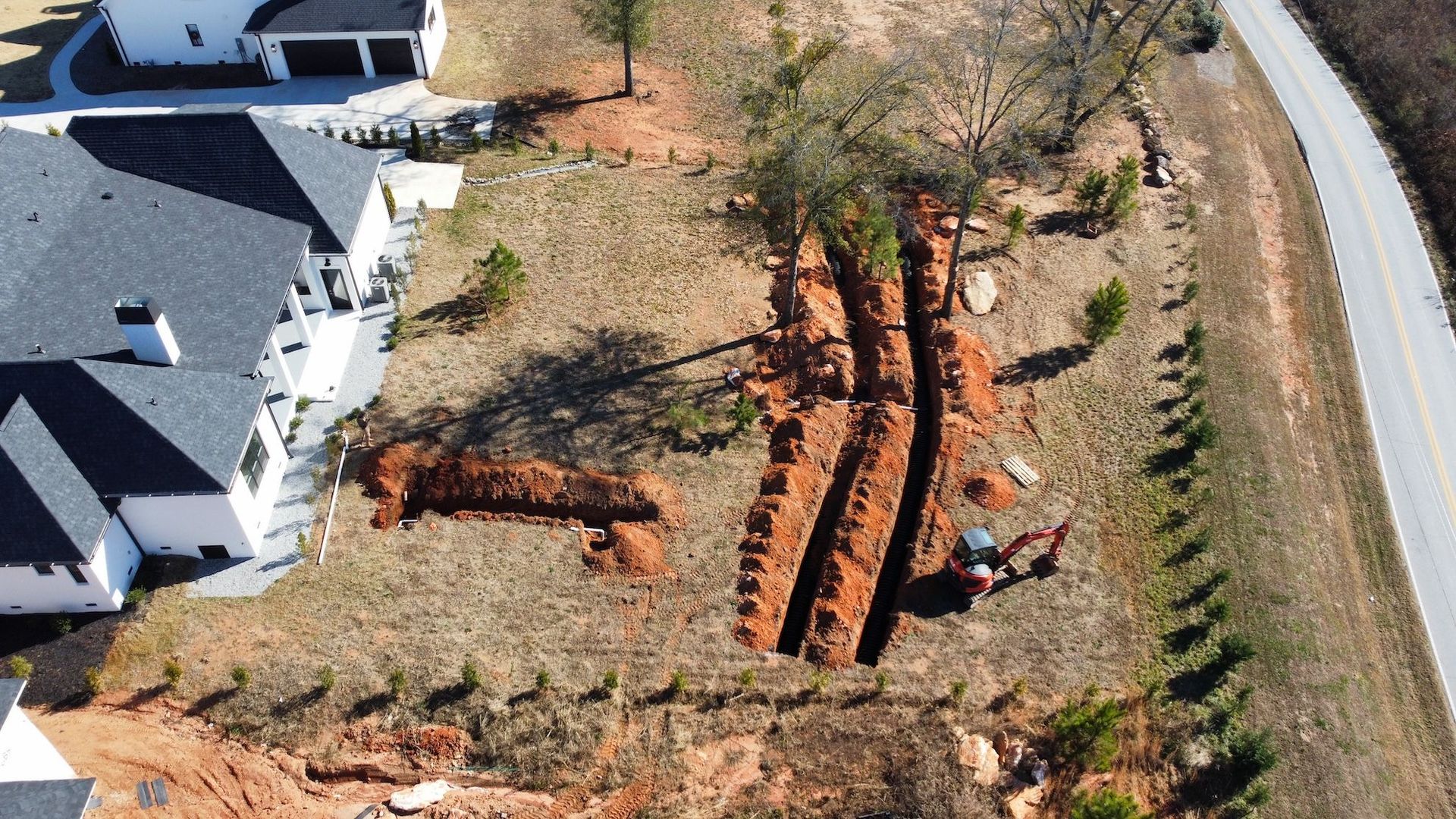 An aerial view of a residential backyard with long, deep trenches dug into the red dirt near a new house and road.