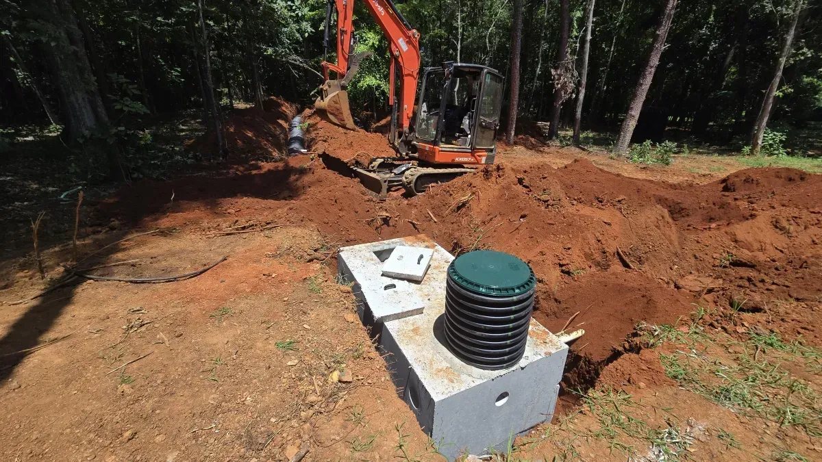 An orange excavator digs in a dirt lot next to a partially buried concrete septic tank with a green access lid.