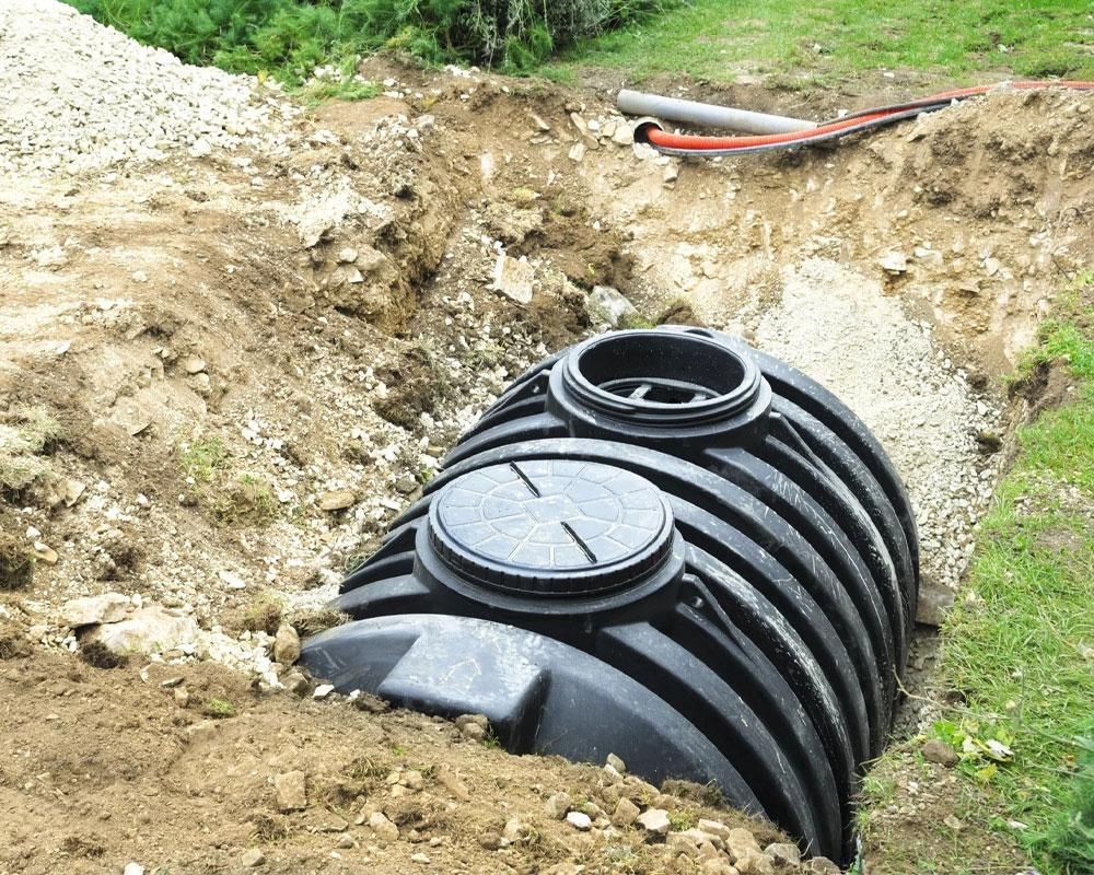 A black plastic septic tank sits in an excavated dirt trench in a grassy area, awaiting installation.