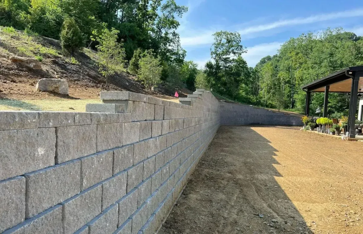 A tan stone retaining wall runs along a dirt yard next to a covered patio with trees in the background.