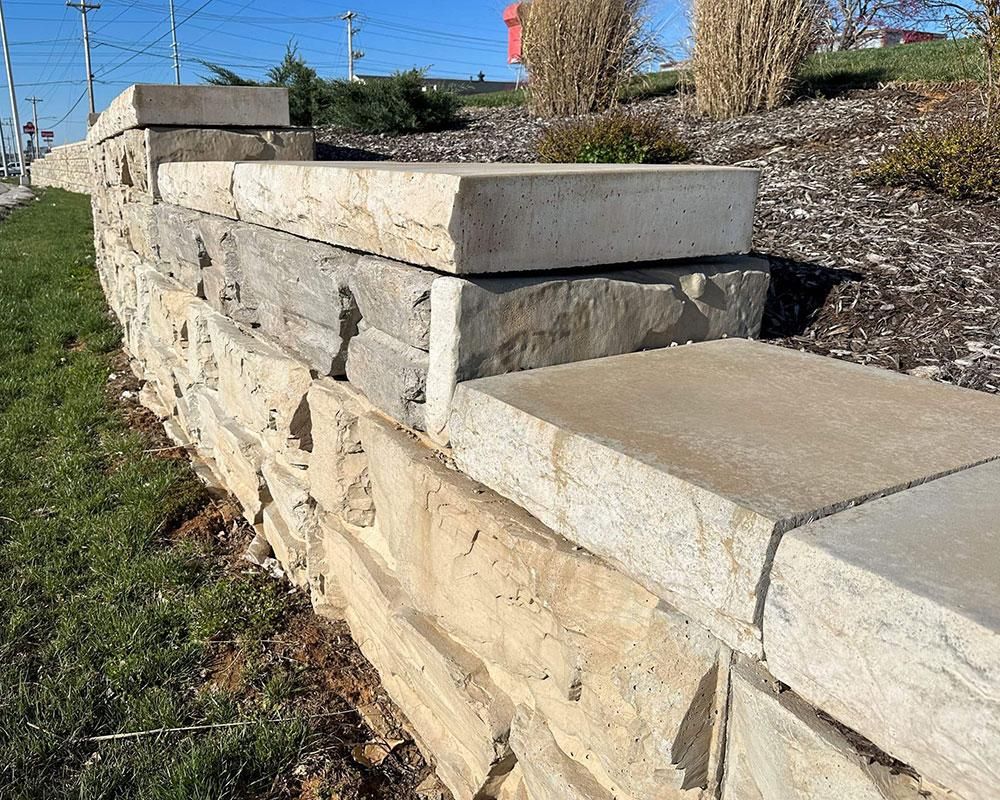 A low retaining wall made of stacked, light-colored natural stone slabs next to a grassy lawn and a mulched slope.