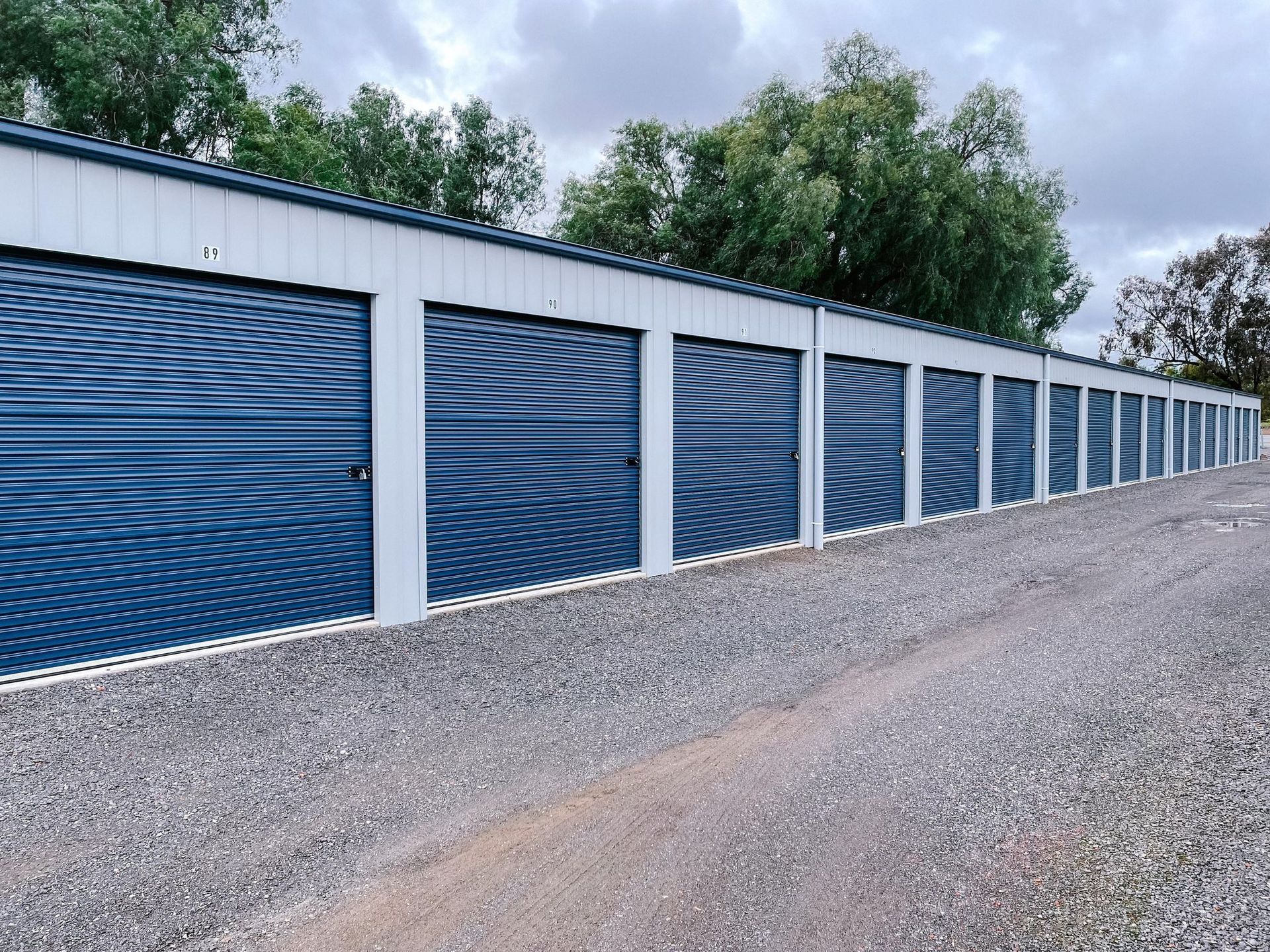 A row of blue garage doors are lined up on a gravel road.