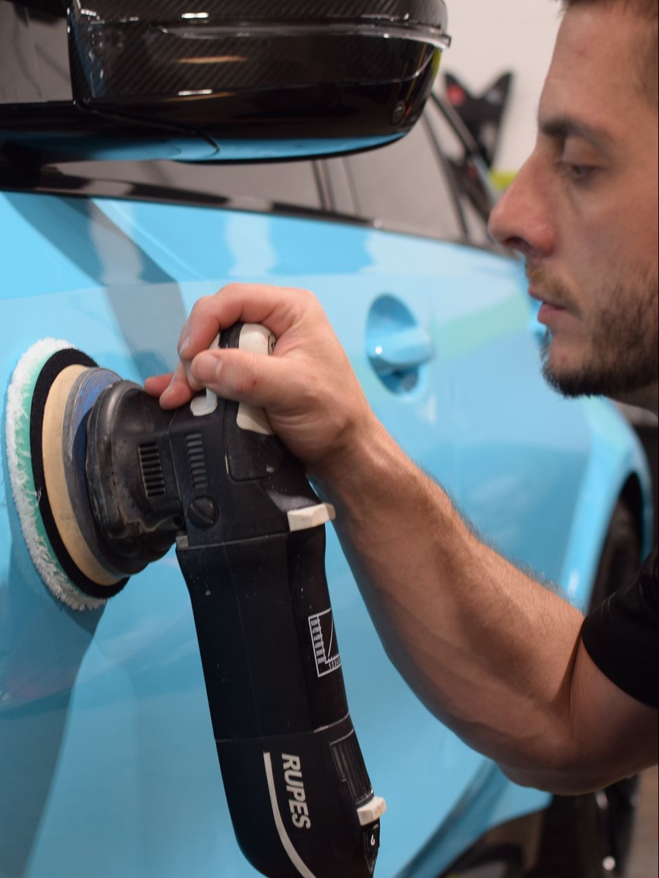 A man is polishing a blue car with a machine made by rupes
