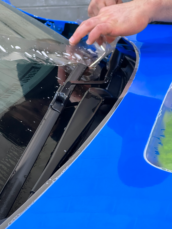 A person is applying a clear film to the windshield of a blue car