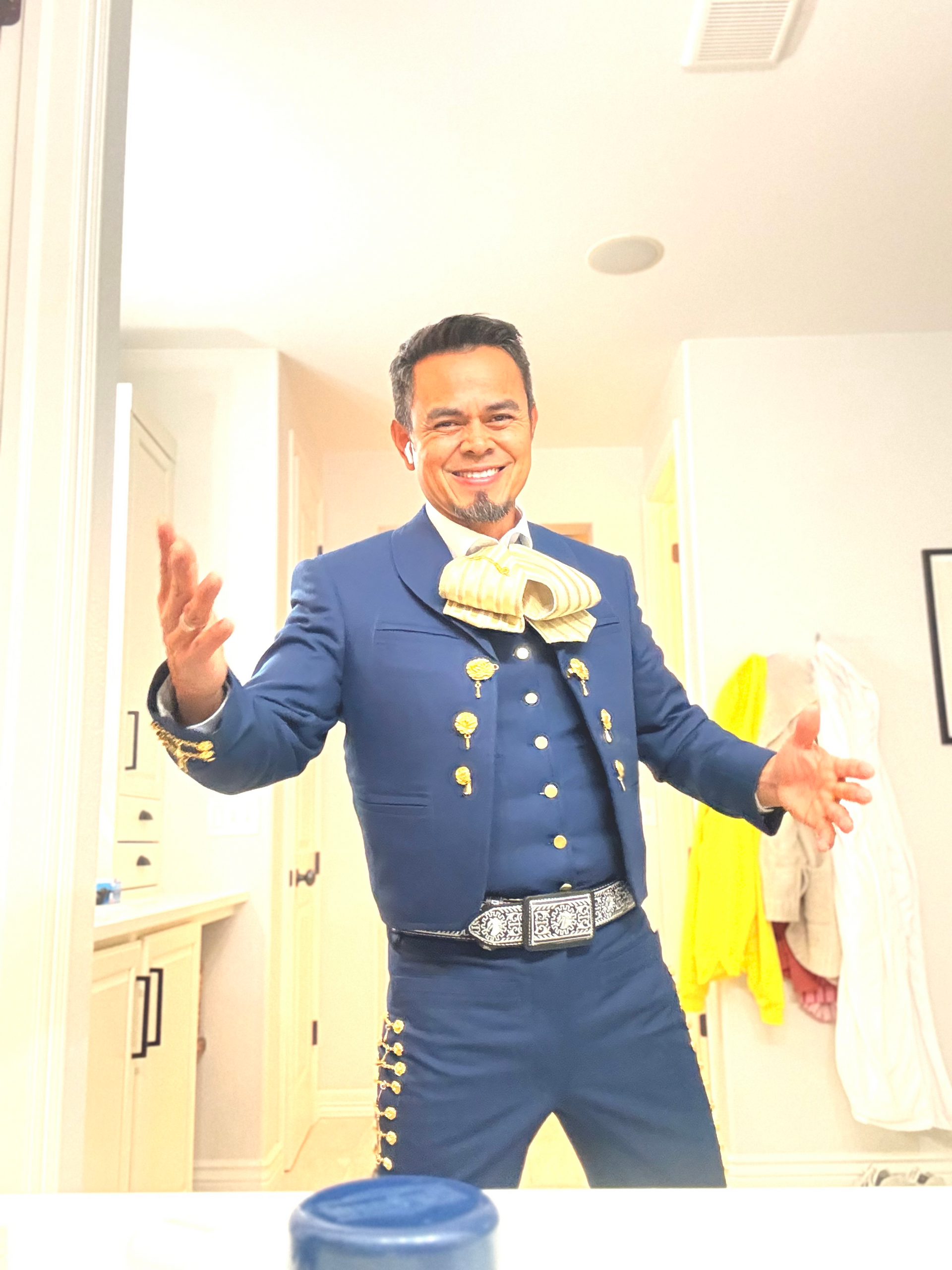 Man in a blue mariachi suit with arms outstretched, smiling. Indoors, bright room.