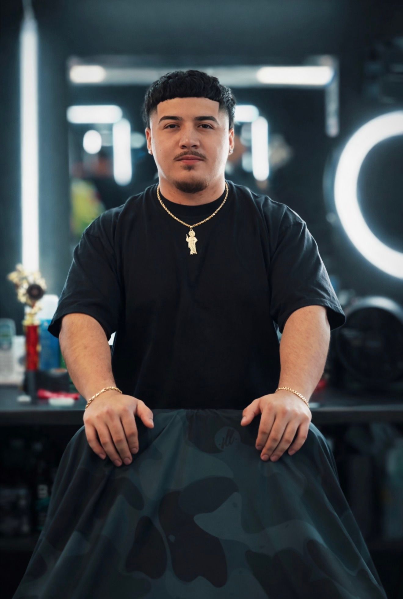 Barber in black shirt stands, hands on cape, in front of mirror with ring light in a shop.