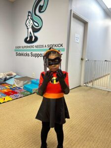 A little girl in a superhero costume is standing in a hallway.