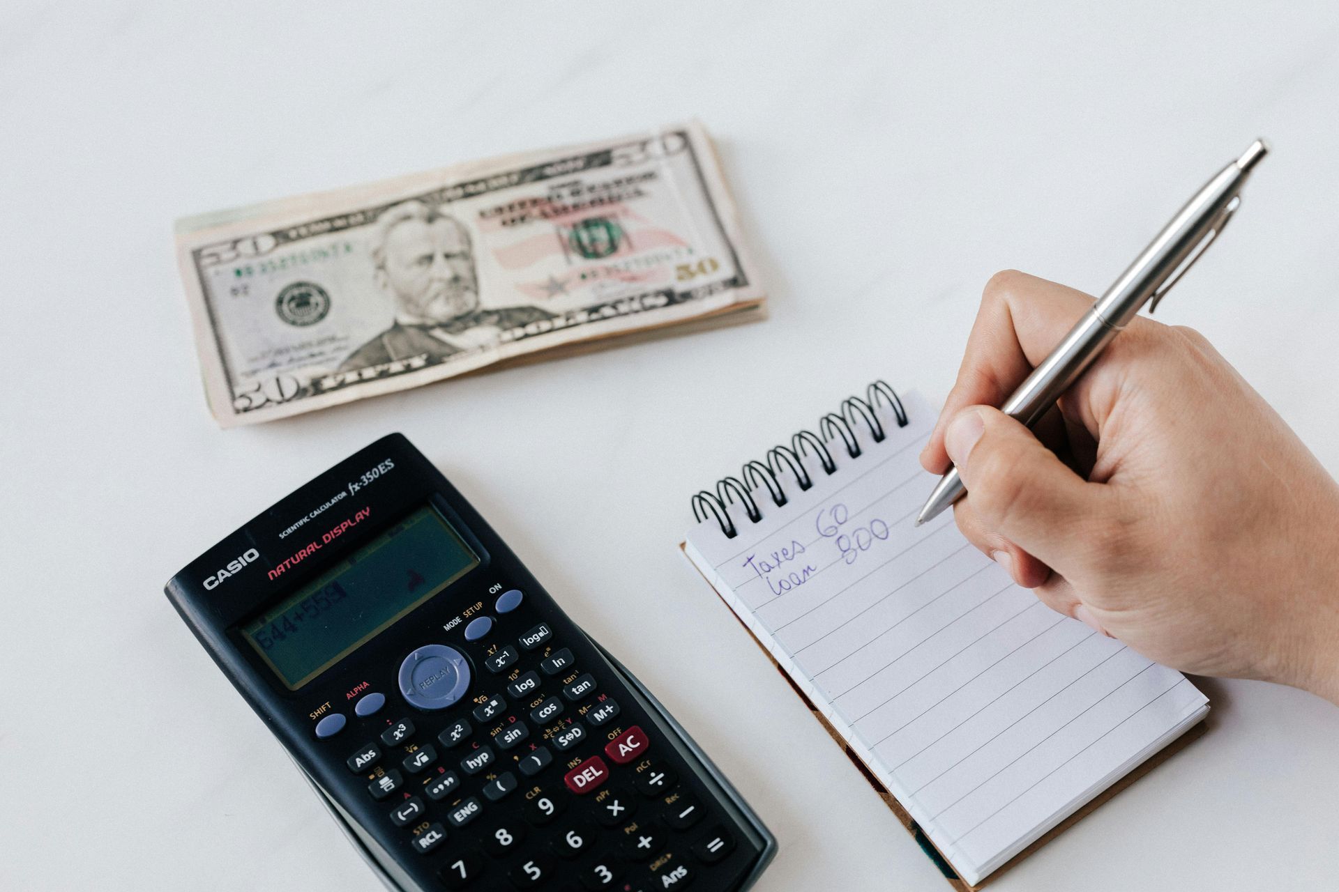 Money, calculator, and notepad on a white surface; hand writing.