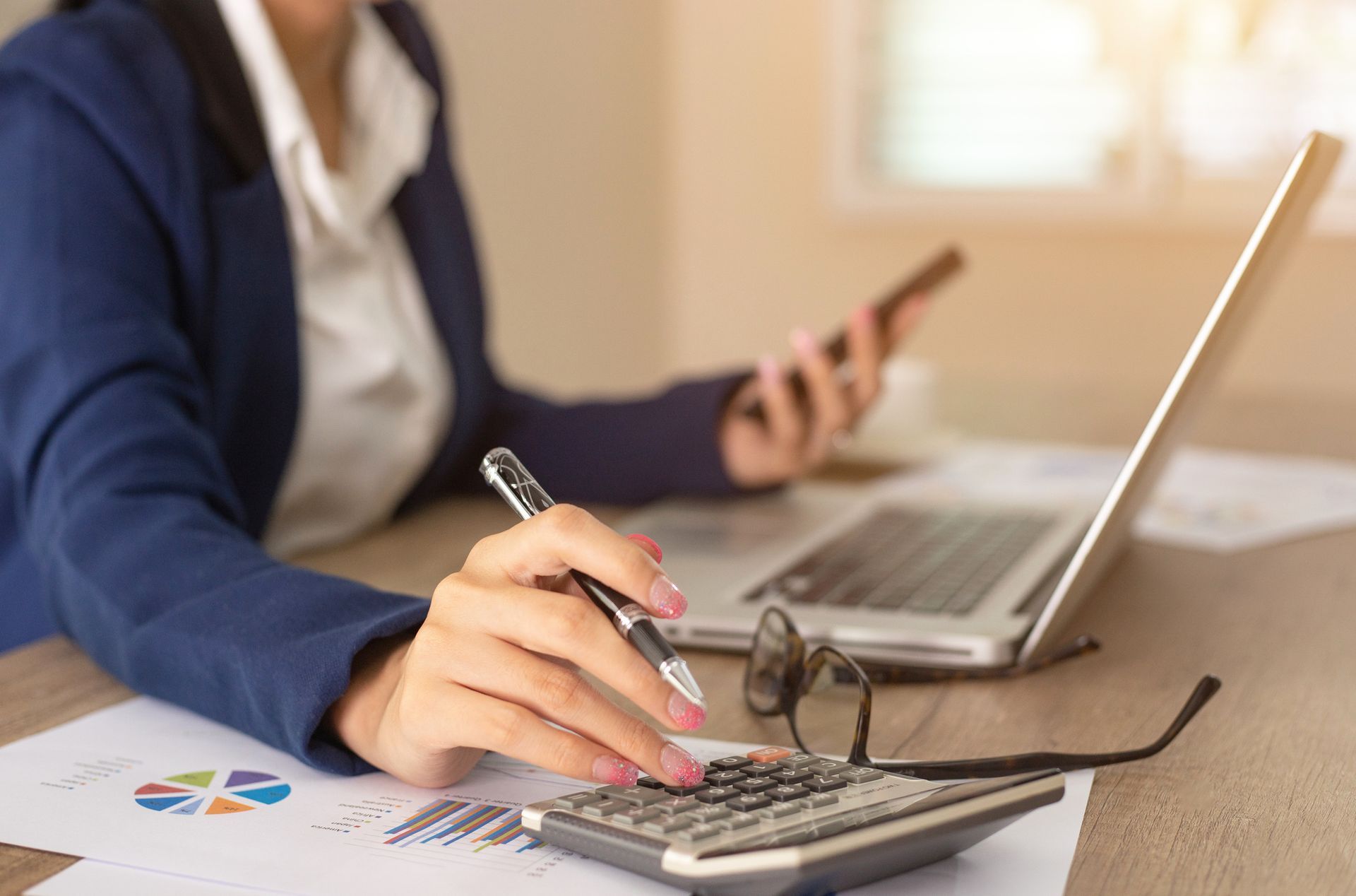A woman is sitting at a desk using a calculator and a laptop computer.