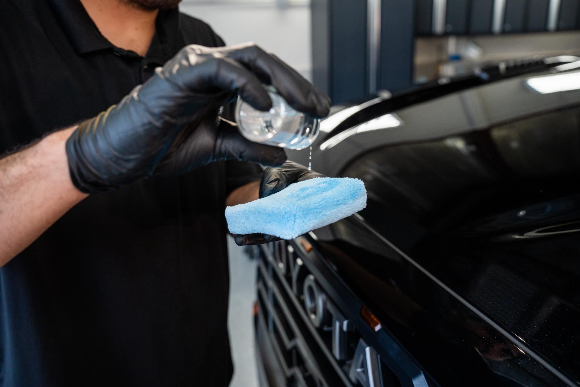 Ceramic Coating - A man wearing black gloves is polishing a black car with a blue sponge .