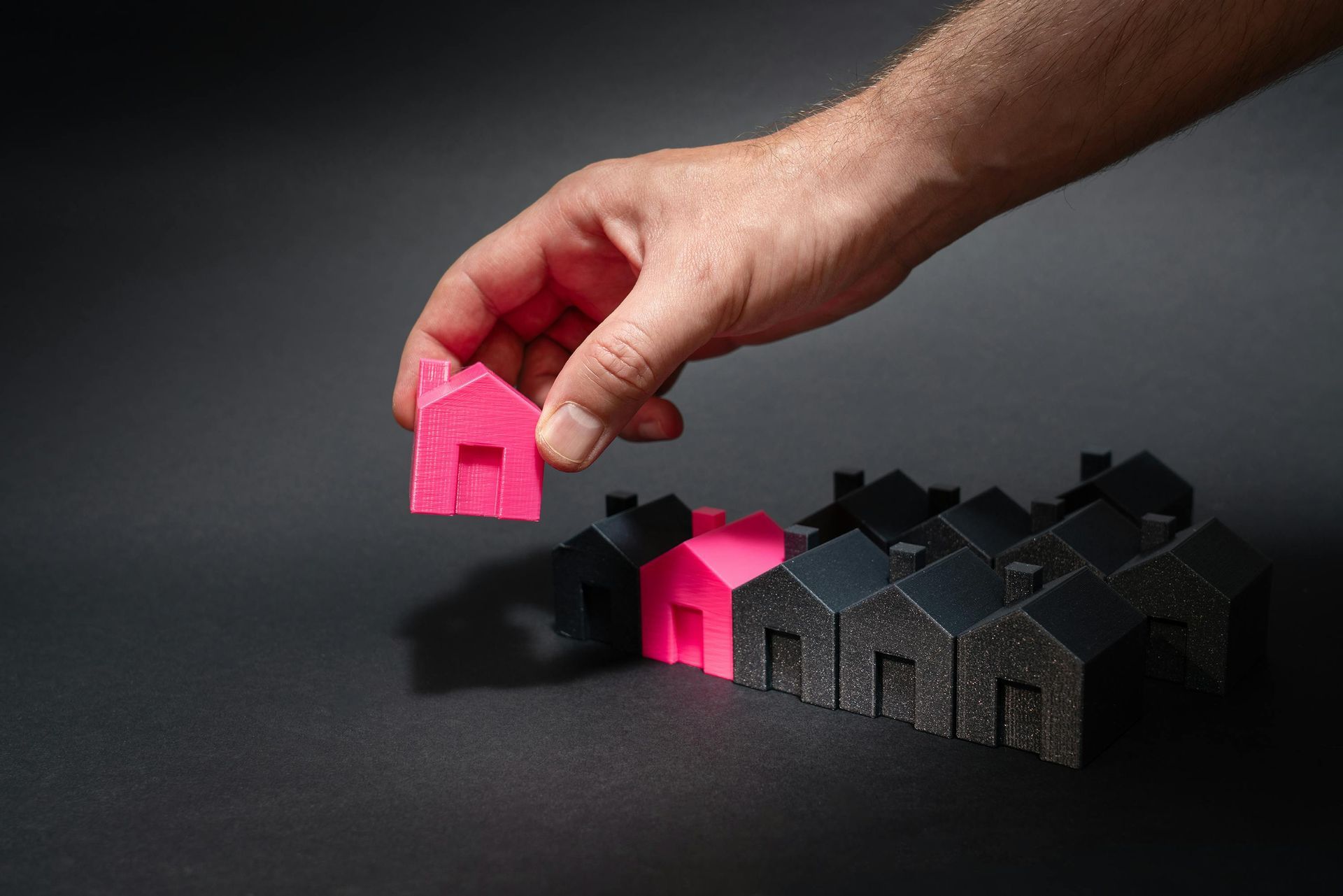 Hand placing a pink toy house beside a row of black toy houses on a dark surface