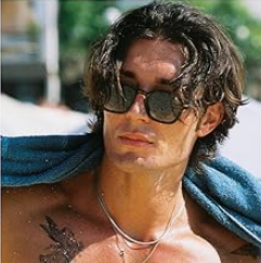 Man with sunglasses, wet hair, bare chest, a towel, and a silver necklace; beach scene.