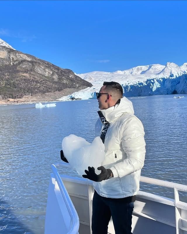 Una persona con chaqueta blanca y gafas de sol sostiene un gran trozo de hielo en un barco cerca de un glaciar y montañas.