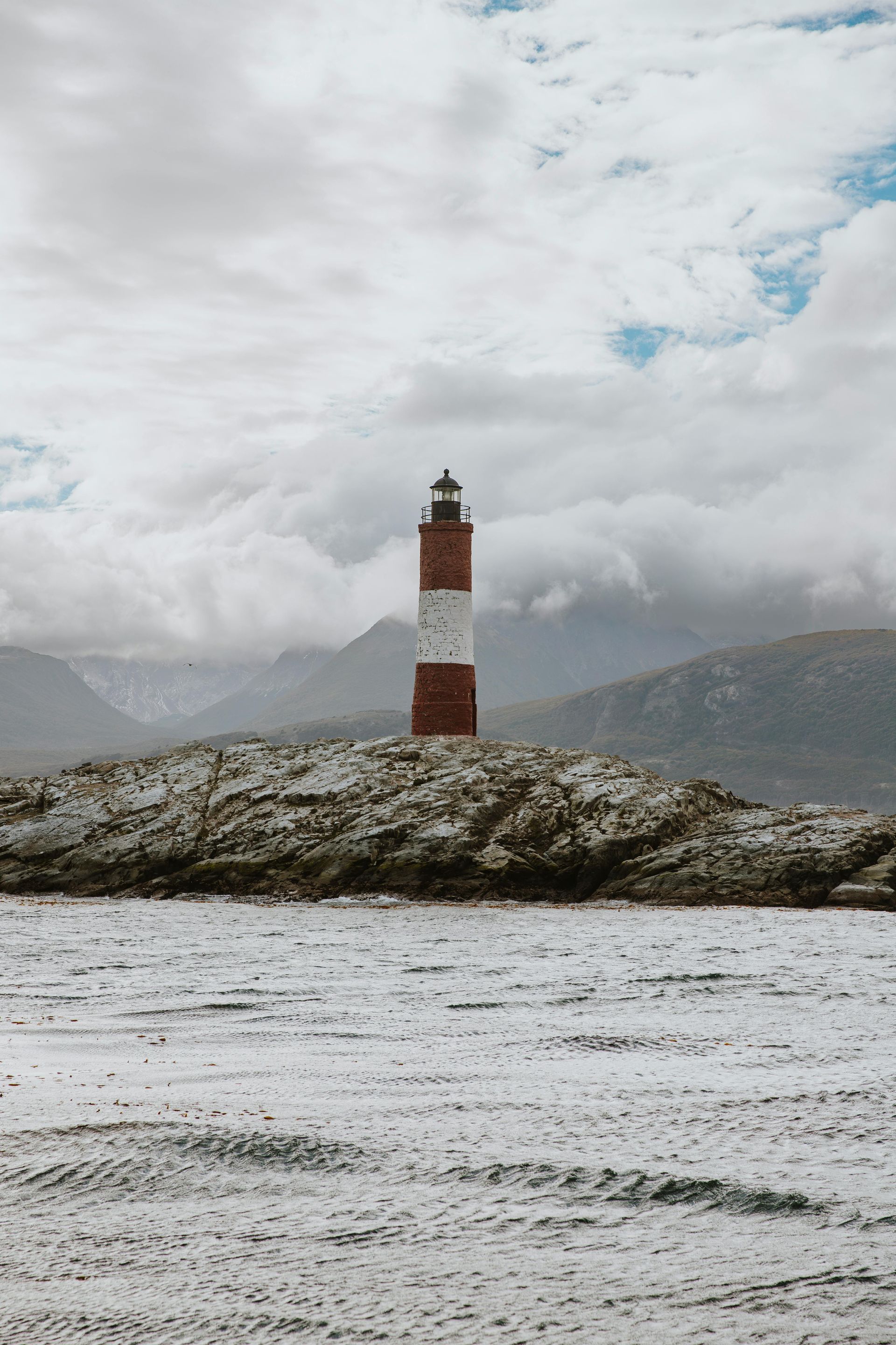 Un faro rojo y blanco se alza sobre un promontorio rocoso cubierto de nieve, con un telón de fondo de montañas nubosas.